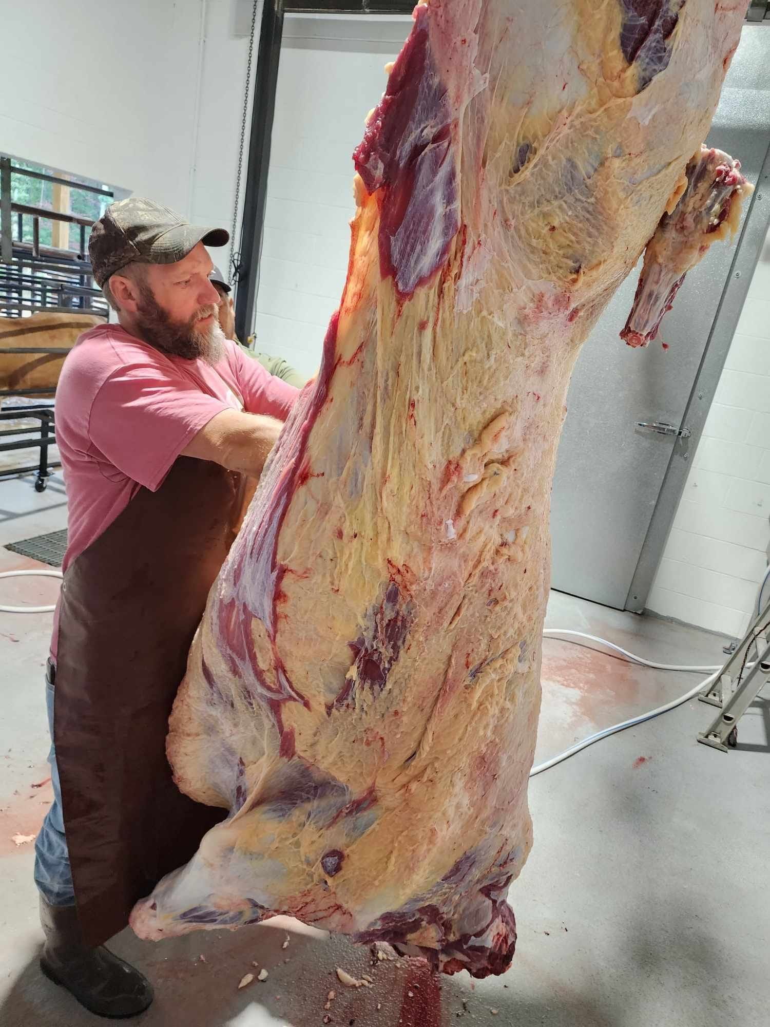 Man butchering a cow carcass. He's wearing an apron and a hat, indoors. Red meat is visible with fat.