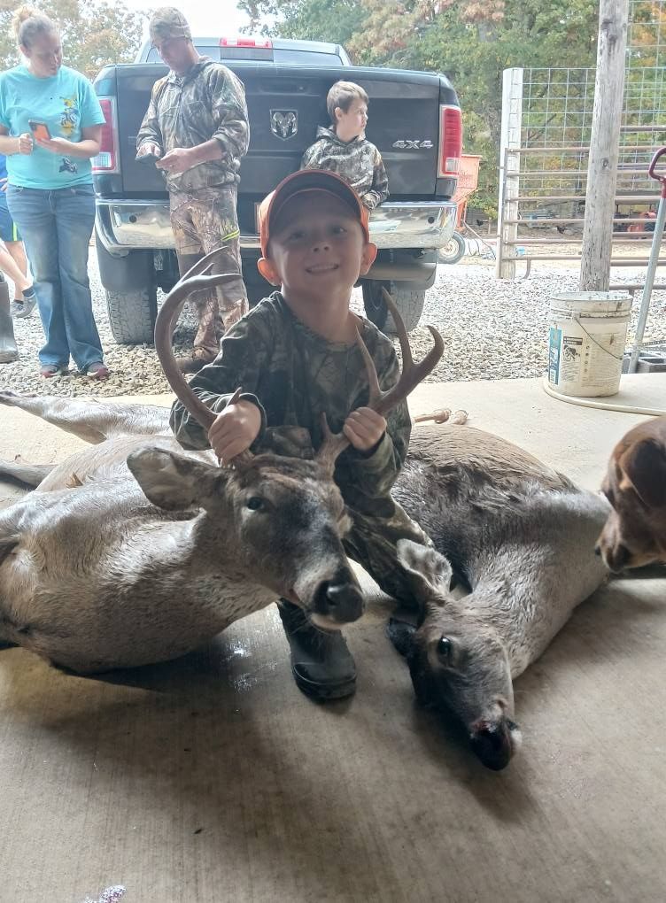 Young boy in camo smiles, holding deer antlers over two deer, in front of a truck.