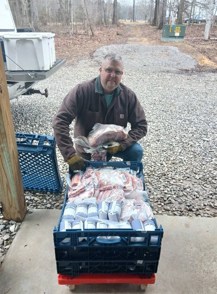 Man with meat packages in a crate, outside.  He smiles, wearing work clothes. Concrete and trees in background.