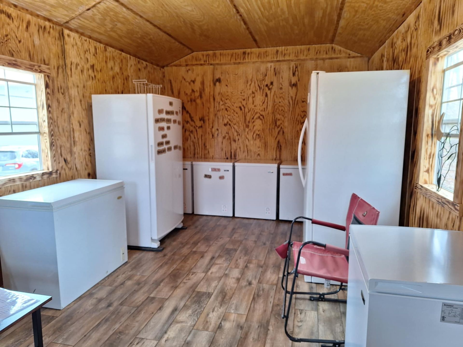 Interior view of a small room with multiple white refrigerators, a freezer, and a chair; wooden walls and floors.