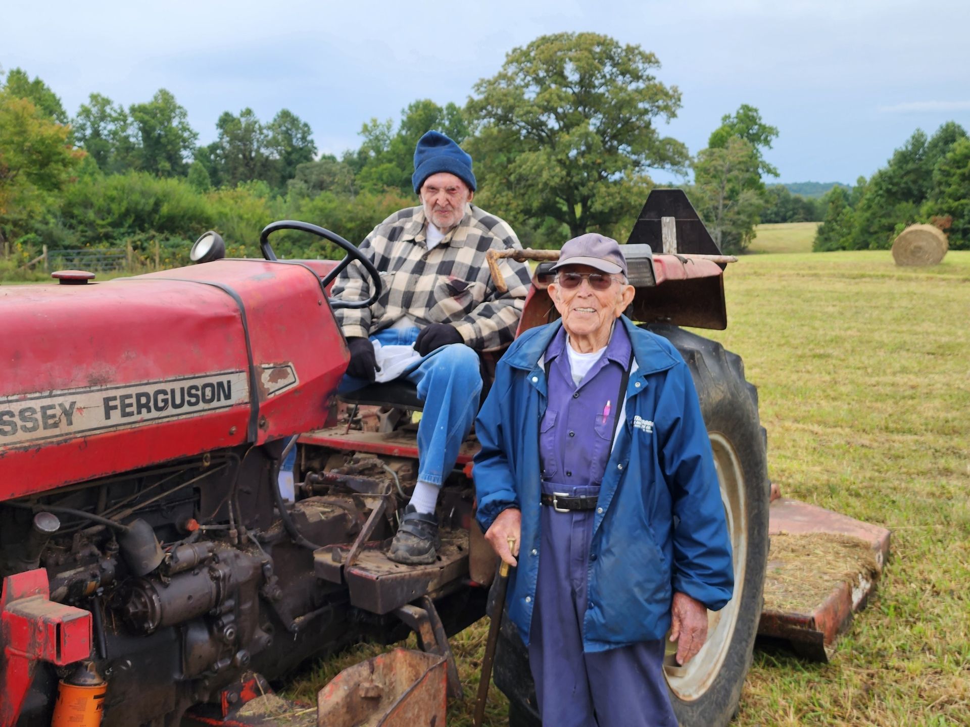 Two men on a red Massey Ferguson tractor in a field. One sits, the other stands, smiling.