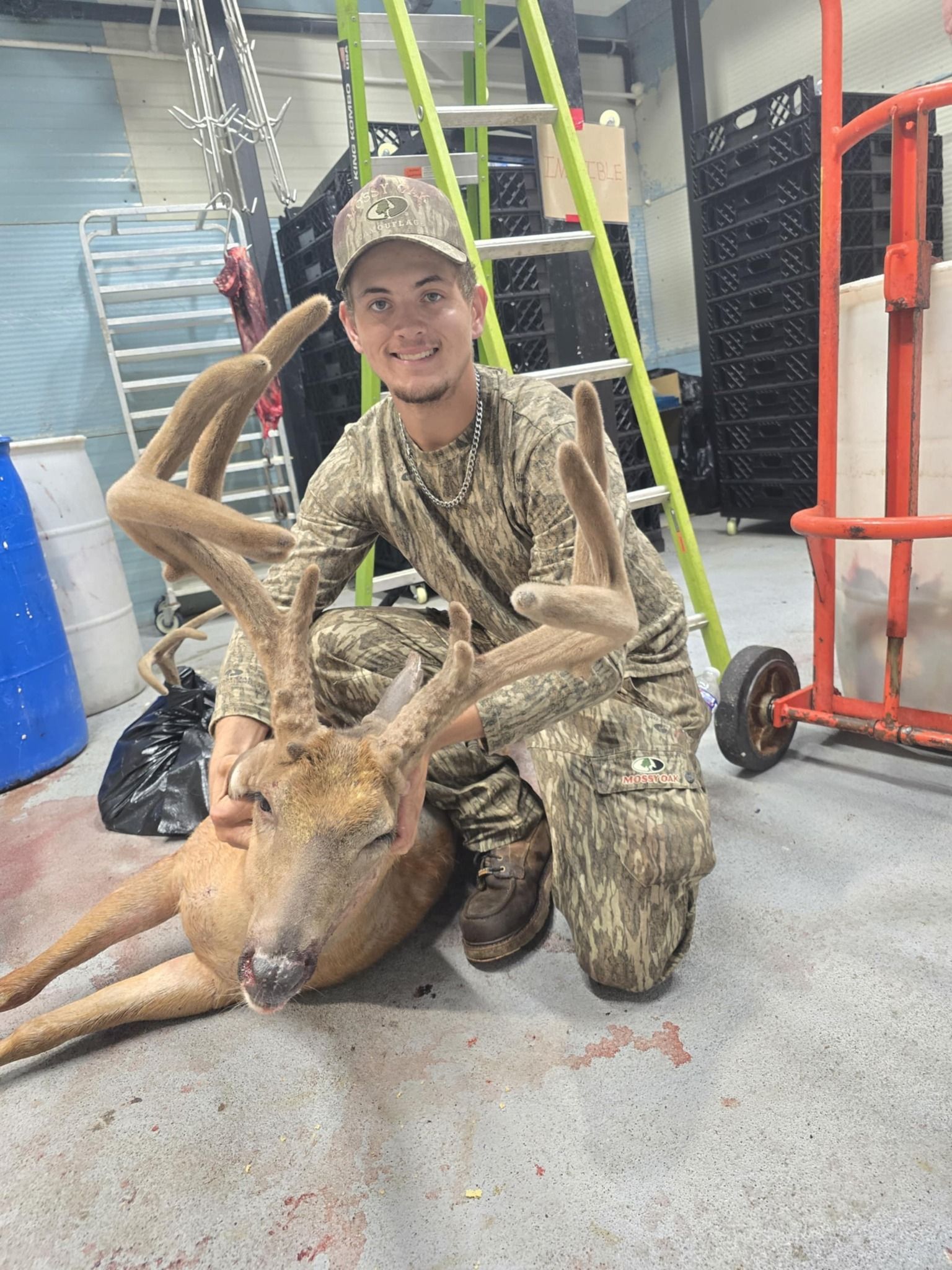 A young man in camouflage kneels next to a harvested deer with large antlers, smiling indoors.