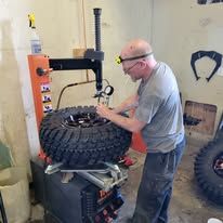 Man in glasses using a tire machine, inside a garage-like space with tools and a large tire.