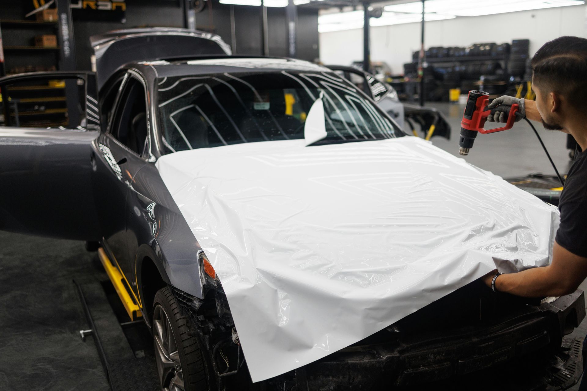 Person applying white vinyl wrap to the hood of a gray car in a shop, using a heat gun.