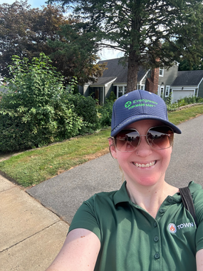 A selfie of a young woman smiling, standing outside of homes in her neighborhood. She is wearing a Town Oil polo shirt, a hat, and sunglasses, and is red-faced from delivering flyers door to door.