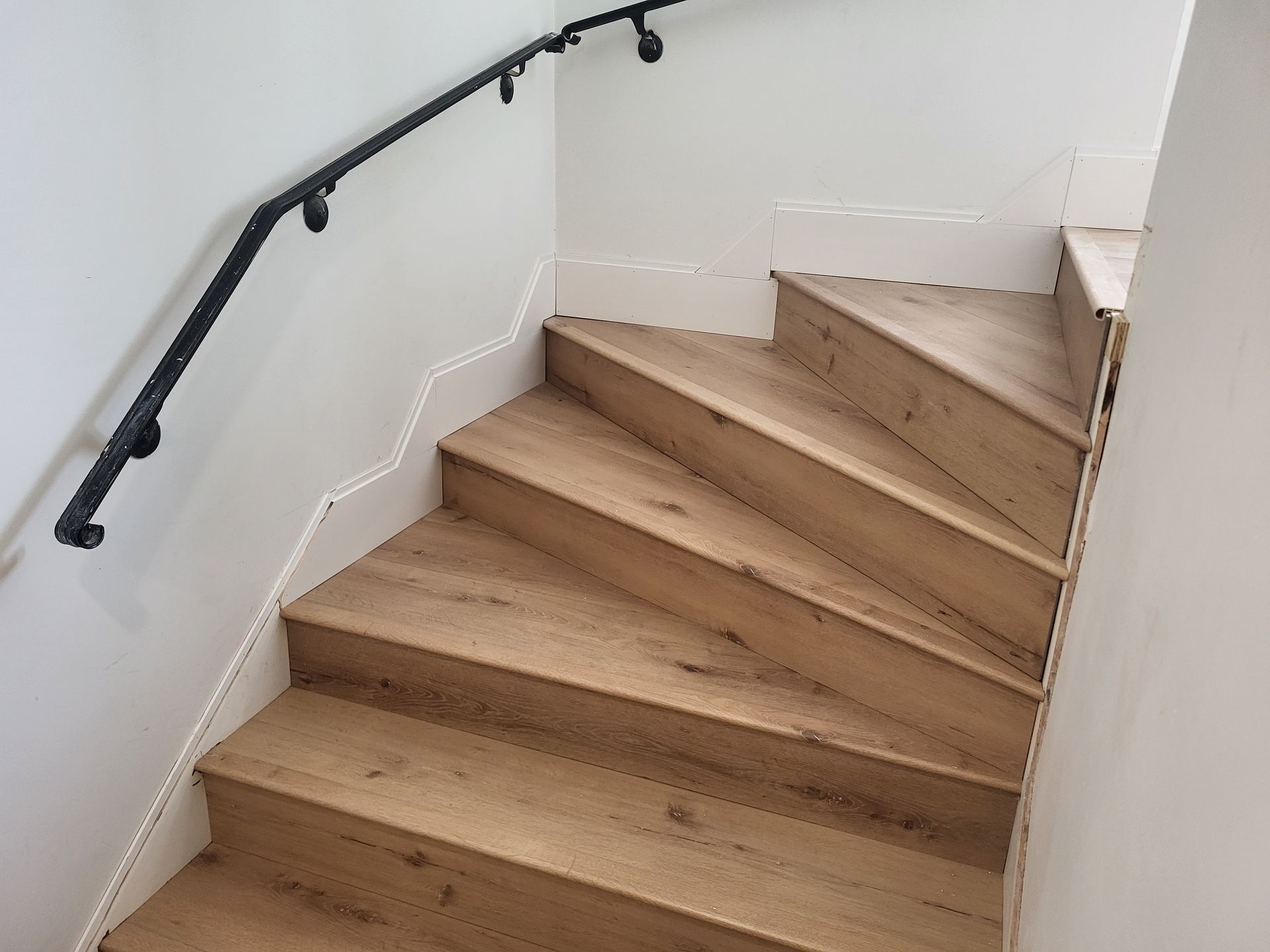 Wooden stairs with a black handrail against a white wall. The steps lead upwards, with a clear, natural wood grain.