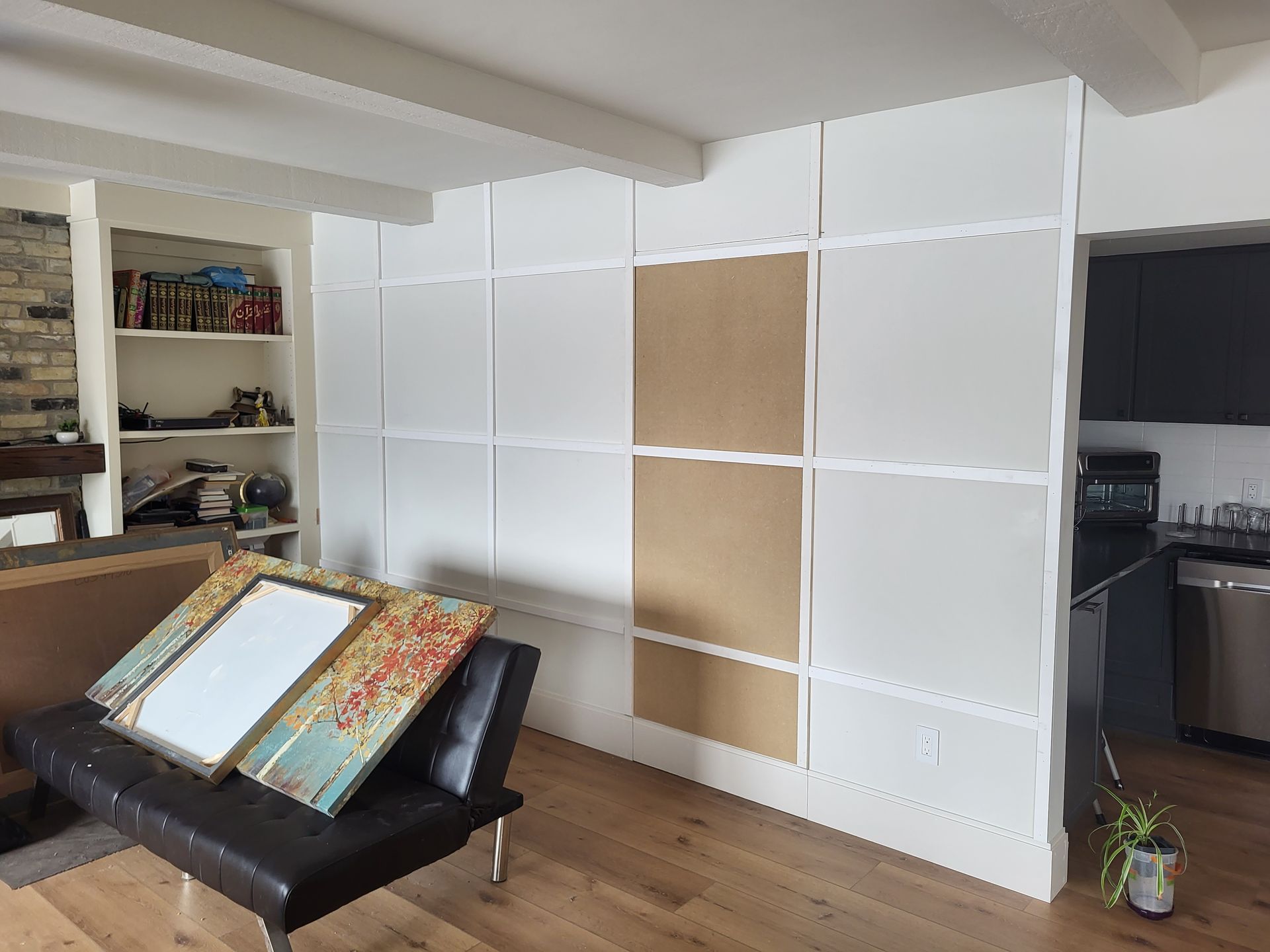 A room with a partially constructed white wall, a black couch, and art leaning against it. A kitchen with dark cabinets is visible on the right.