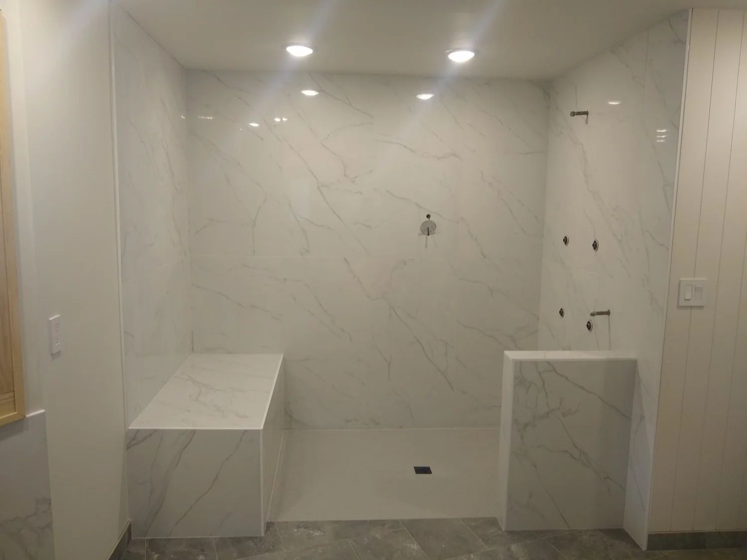 White marble shower with built-in bench and half-wall. Recessed lighting illuminates the space.