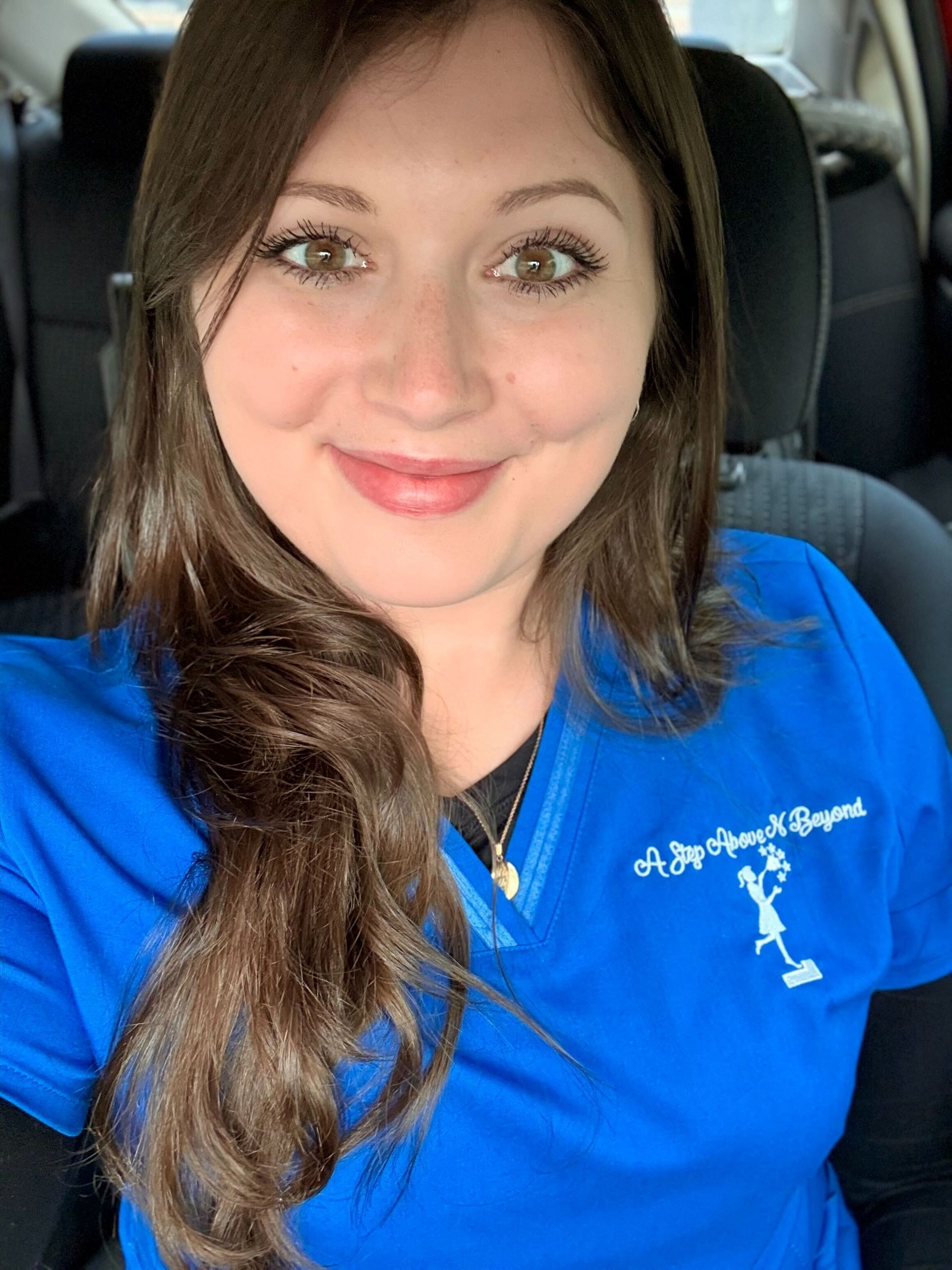 Woman in blue scrubs smiling, in a car. Scrubs have a logo. Brown hair, light skin.