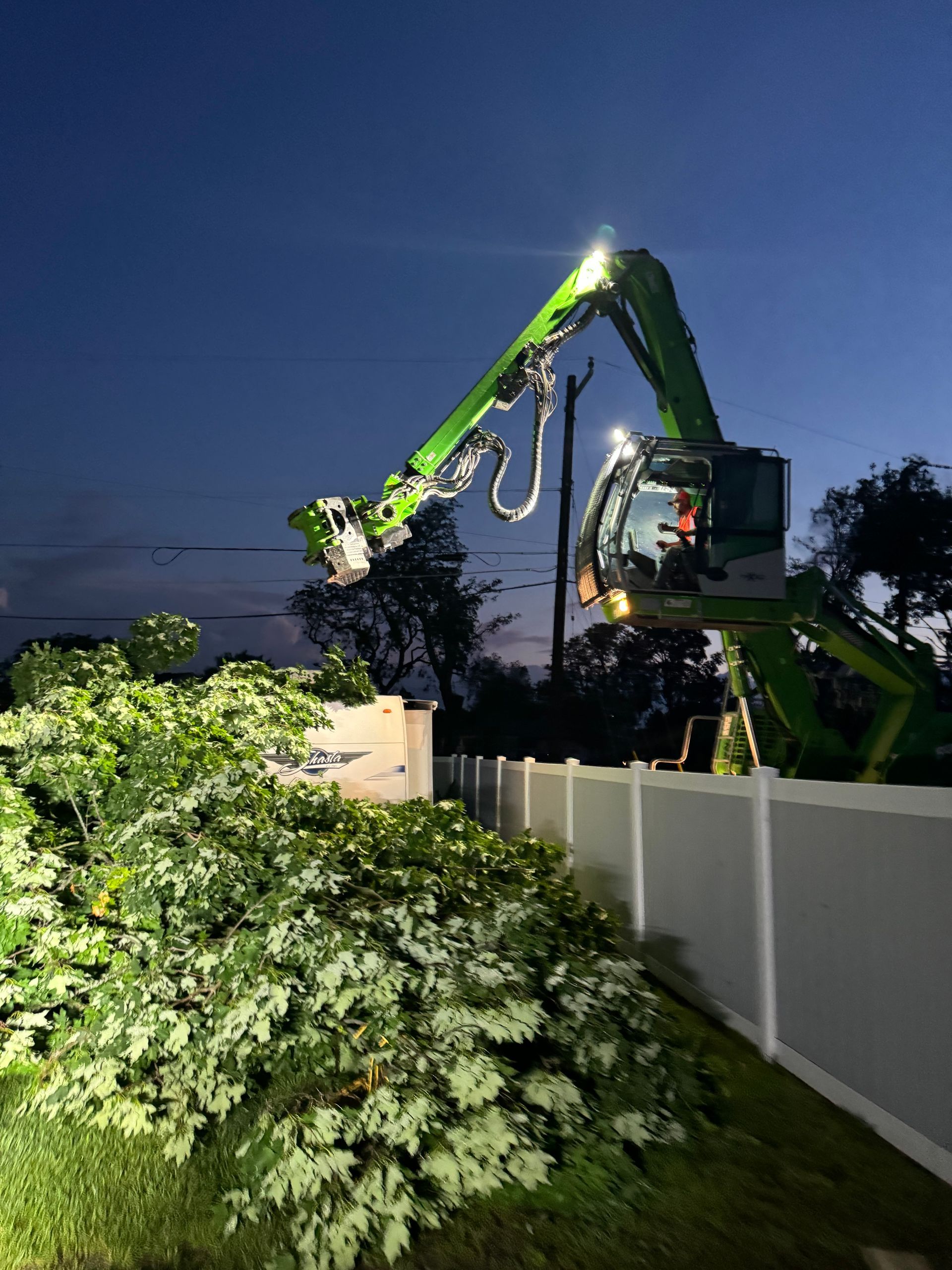 A green crane is cutting a tree in front of a white fence at night.