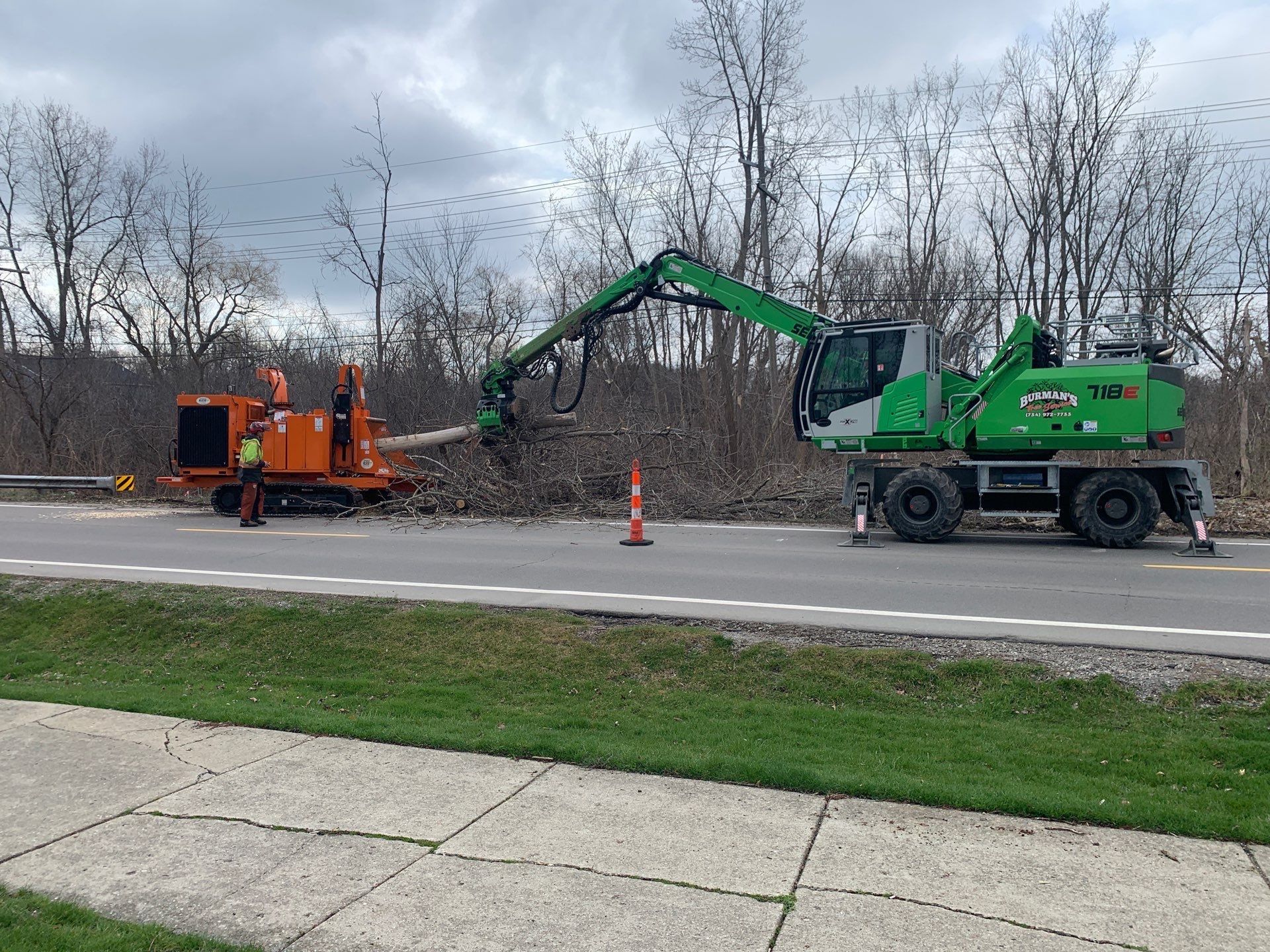A green excavator is cutting down trees on the side of the road.