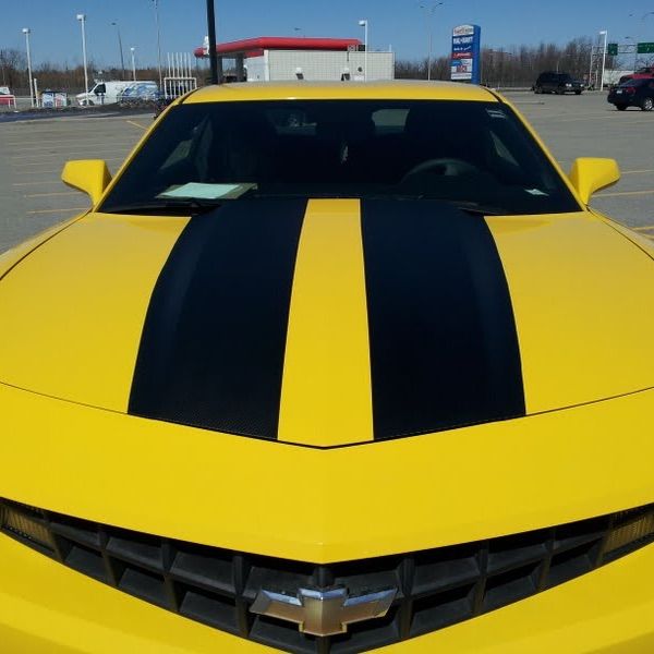 A yellow car with black stripes on the hood is parked in a parking lot