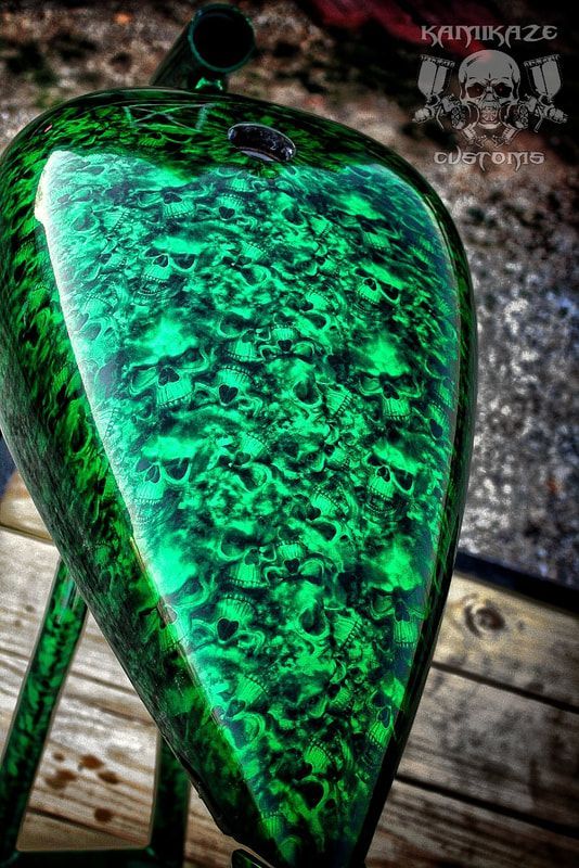 A green motorcycle tank with skulls painted on it is sitting on a wooden table.
