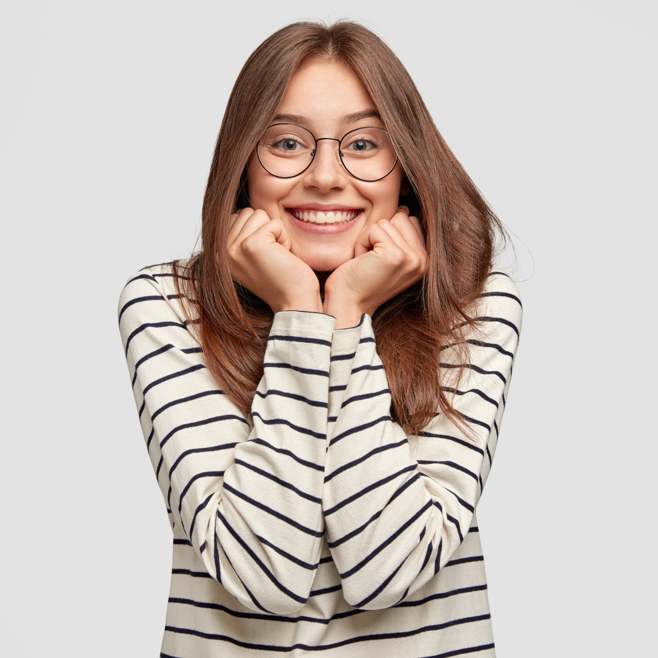 A woman wearing glasses and a striped shirt is smiling with her hands on her face.