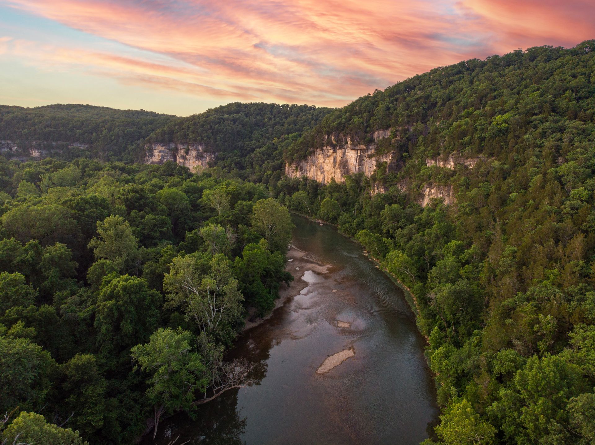 An aerial view of a river surrounded by trees and mountains at sunset.