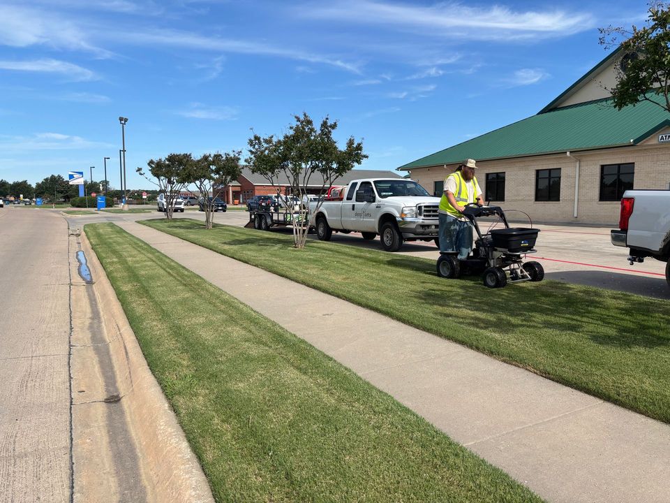 Person on a standing lawn mower next to vehicles on a sidewalk near a building with a green roof.