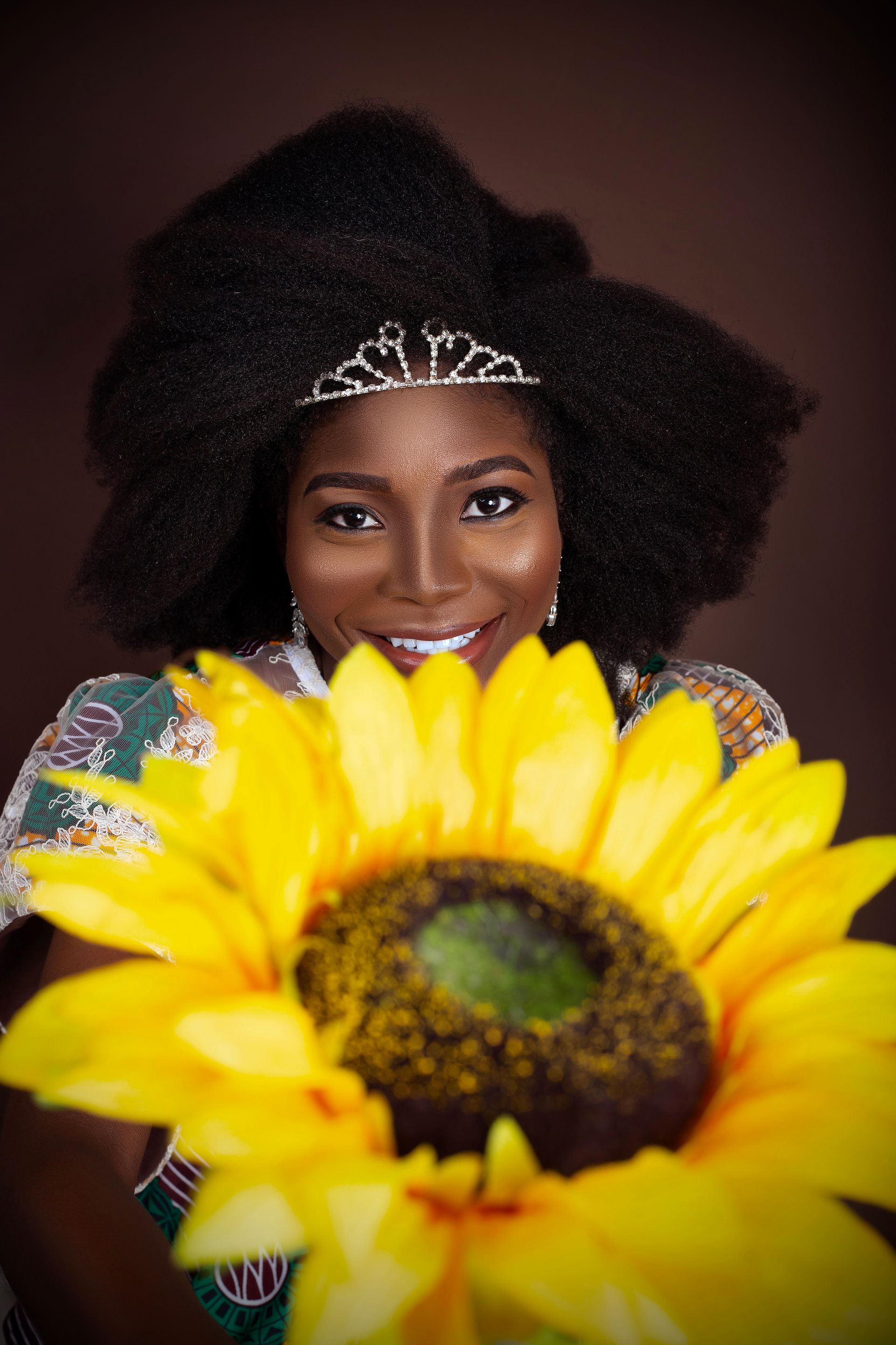 A woman wearing a tiara is holding a sunflower.
