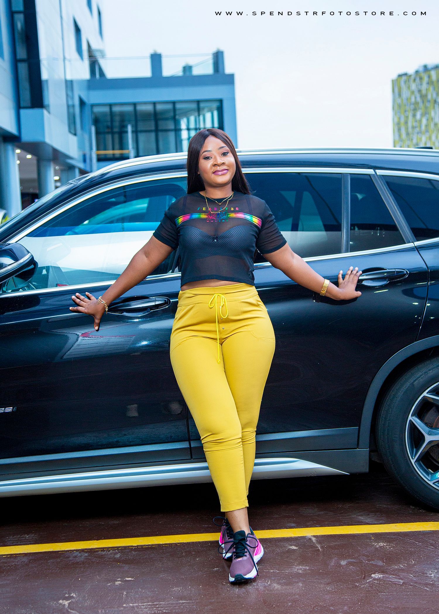A woman in yellow pants is standing next to a black car.