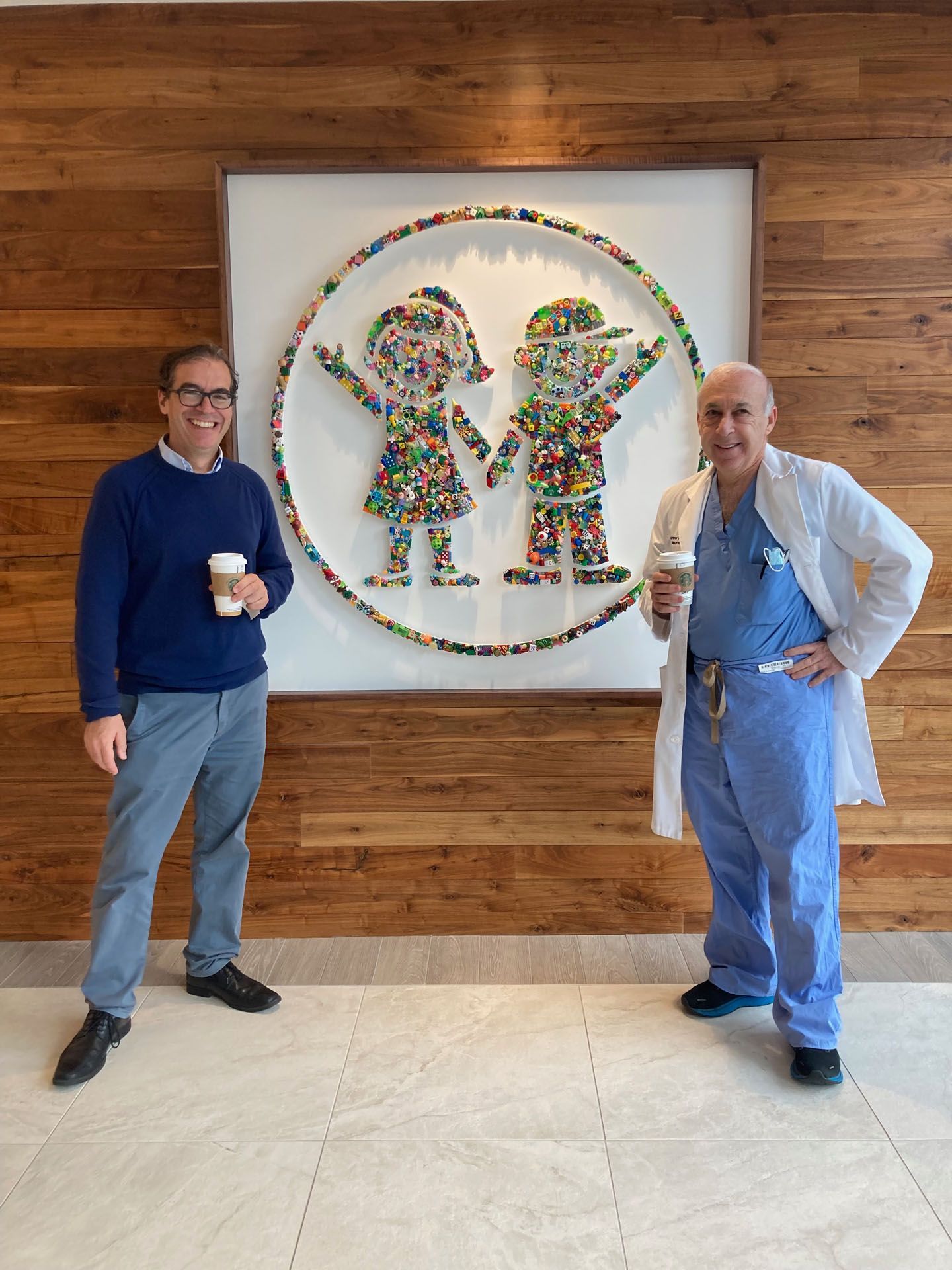 Two men stand beside colorful mosaic art of children. One wears a sweater, the other a lab coat. Both hold coffee cups.