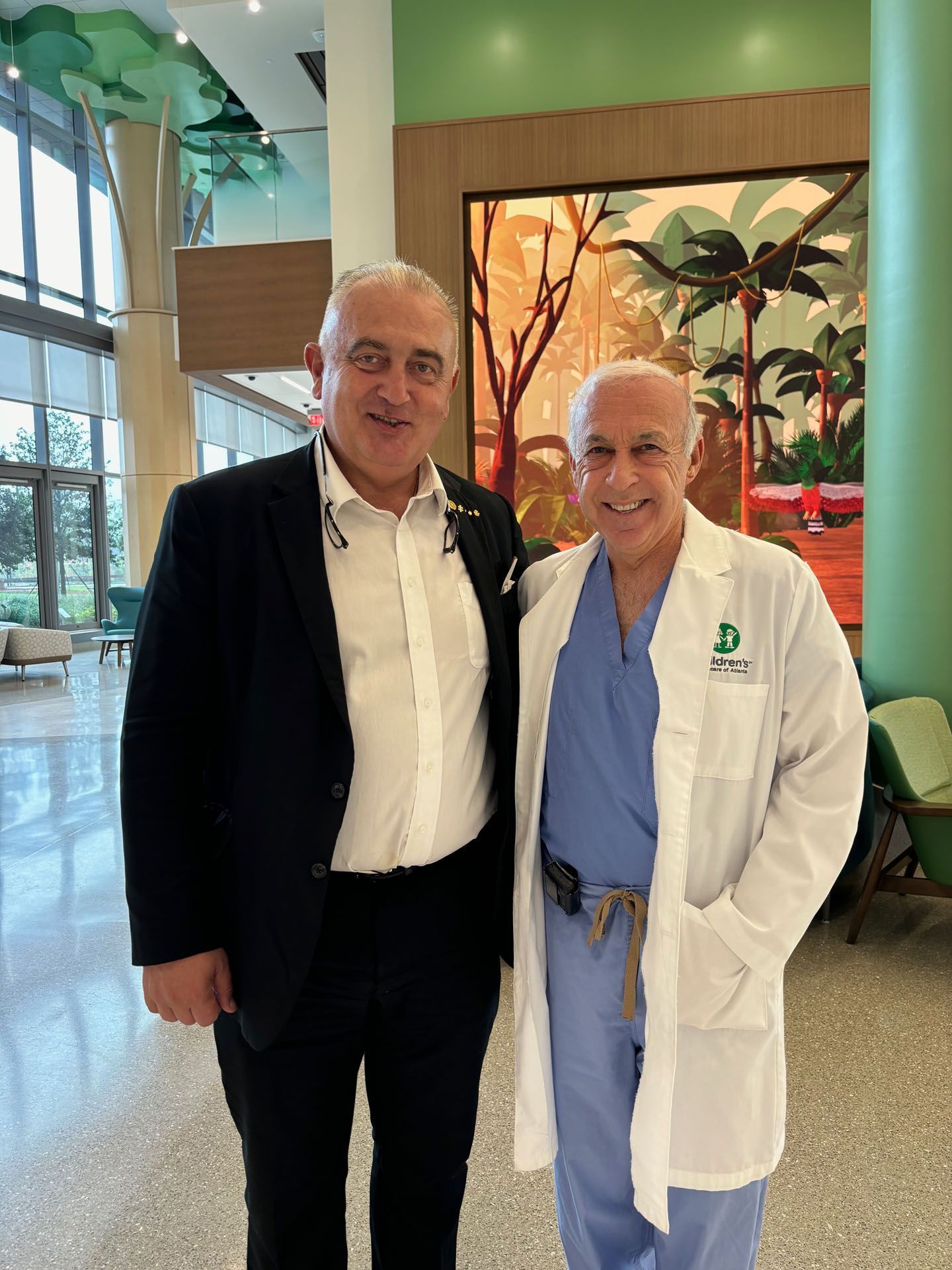 Two men smiling indoors. One in a suit, the other in scrubs and a lab coat, with colorful art in the background.