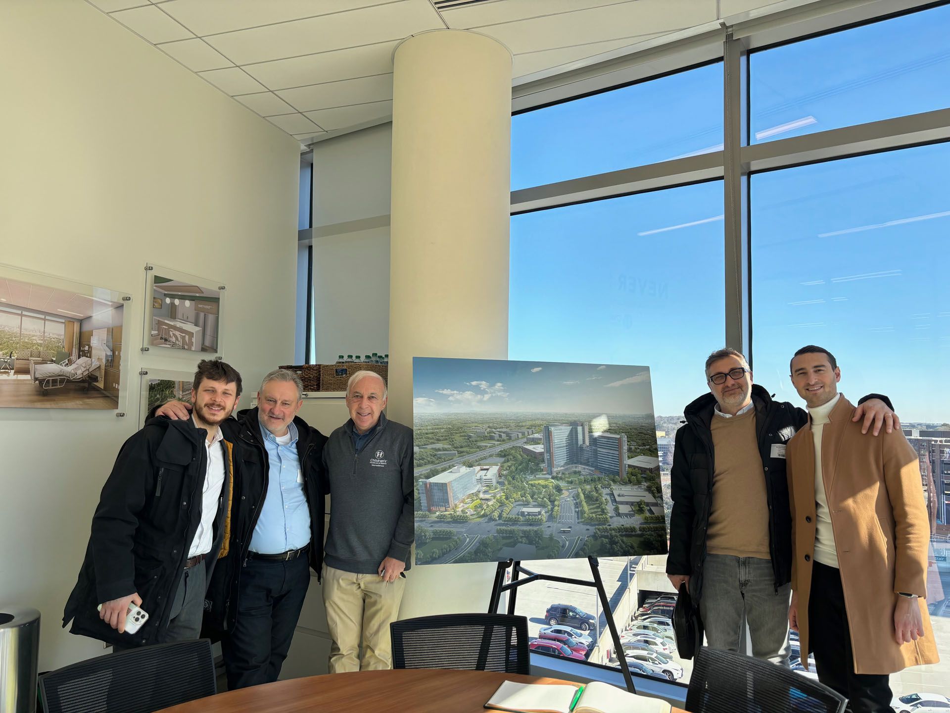 Group of men posing in office with building renderings, sunny day outside window.