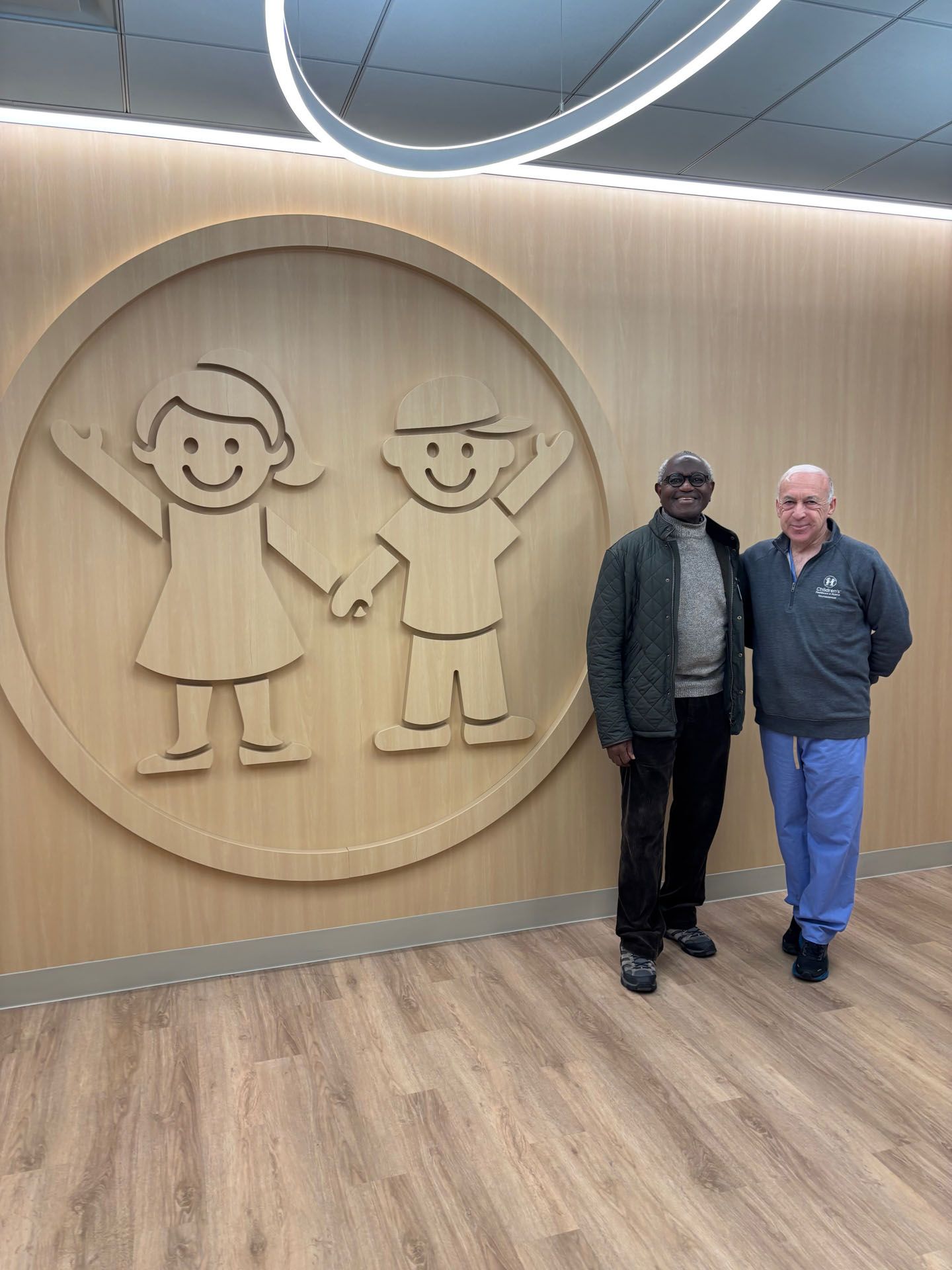 Two men stand by a wooden wall with a circular logo of stick figure children.