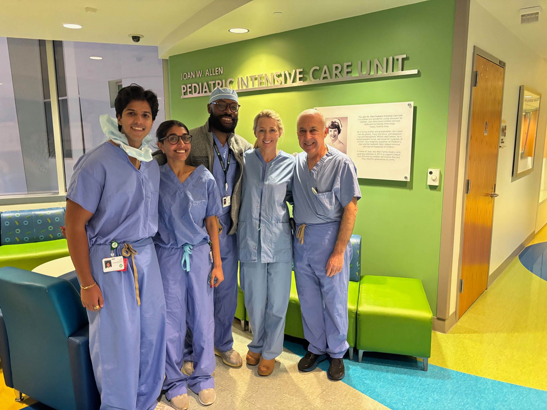 Medical staff pose in front of a Pediatric Intensive Care Unit sign, wearing scrubs.