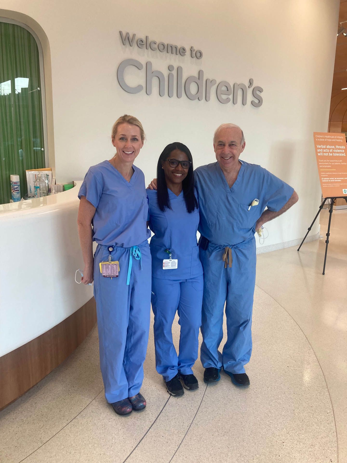 Three healthcare workers in blue scrubs smiling at Children's hospital entrance.
