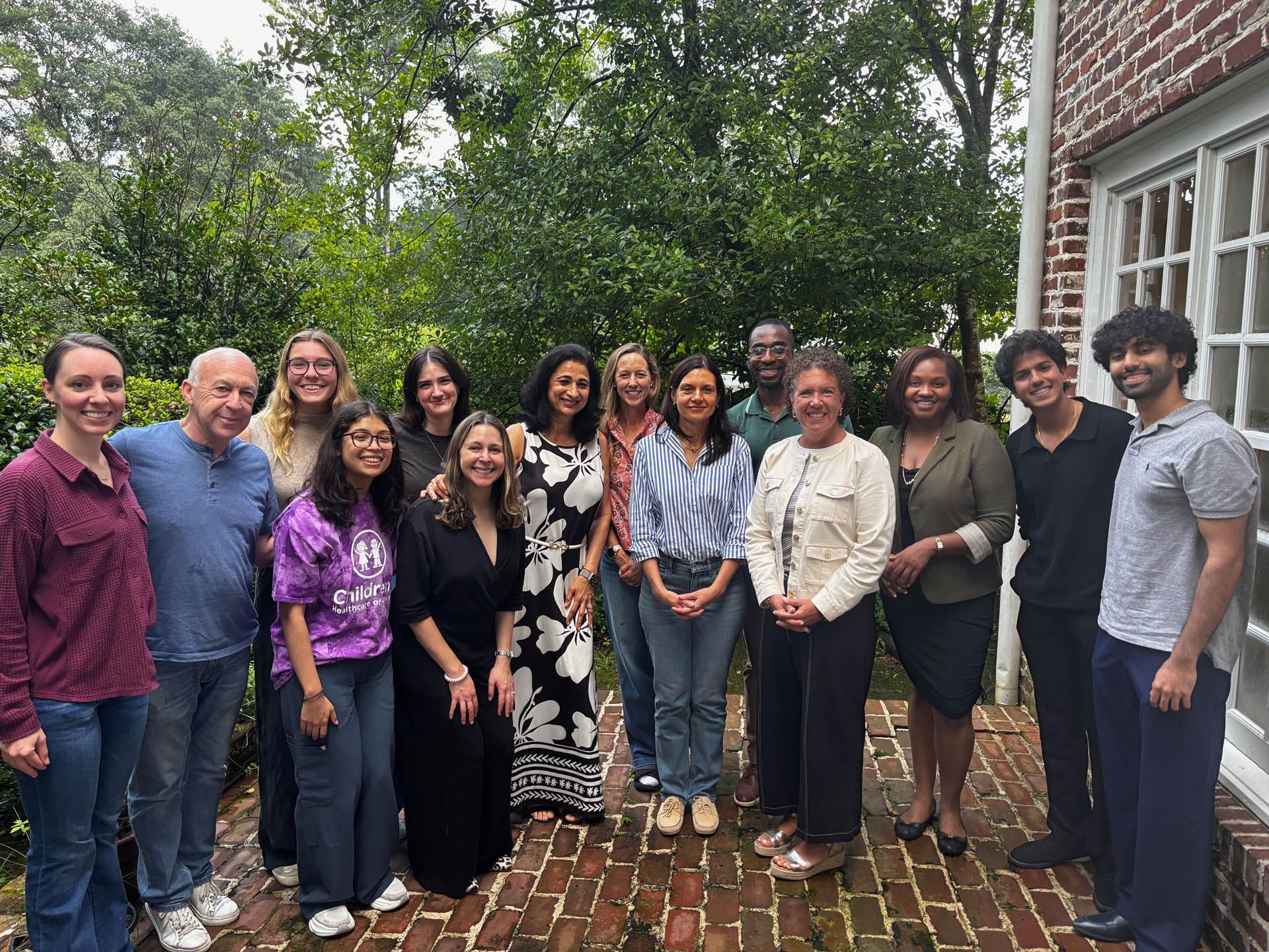Group of diverse people smiling, posing outdoors on a brick patio. Green trees and a white-walled building in the background.