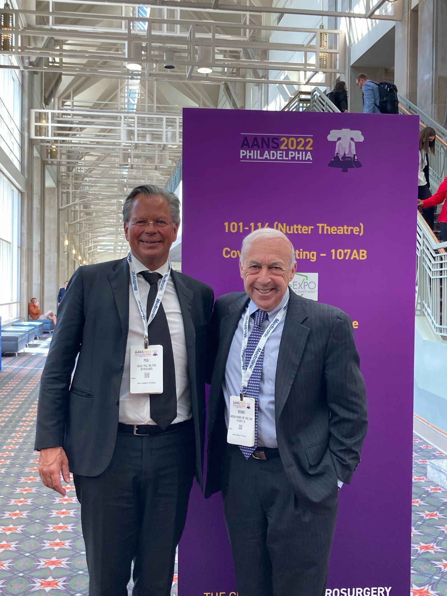 Two men pose by a purple banner at a conference. Both wear suits, smiling. The banner says DANE 2022, Philadelphia.