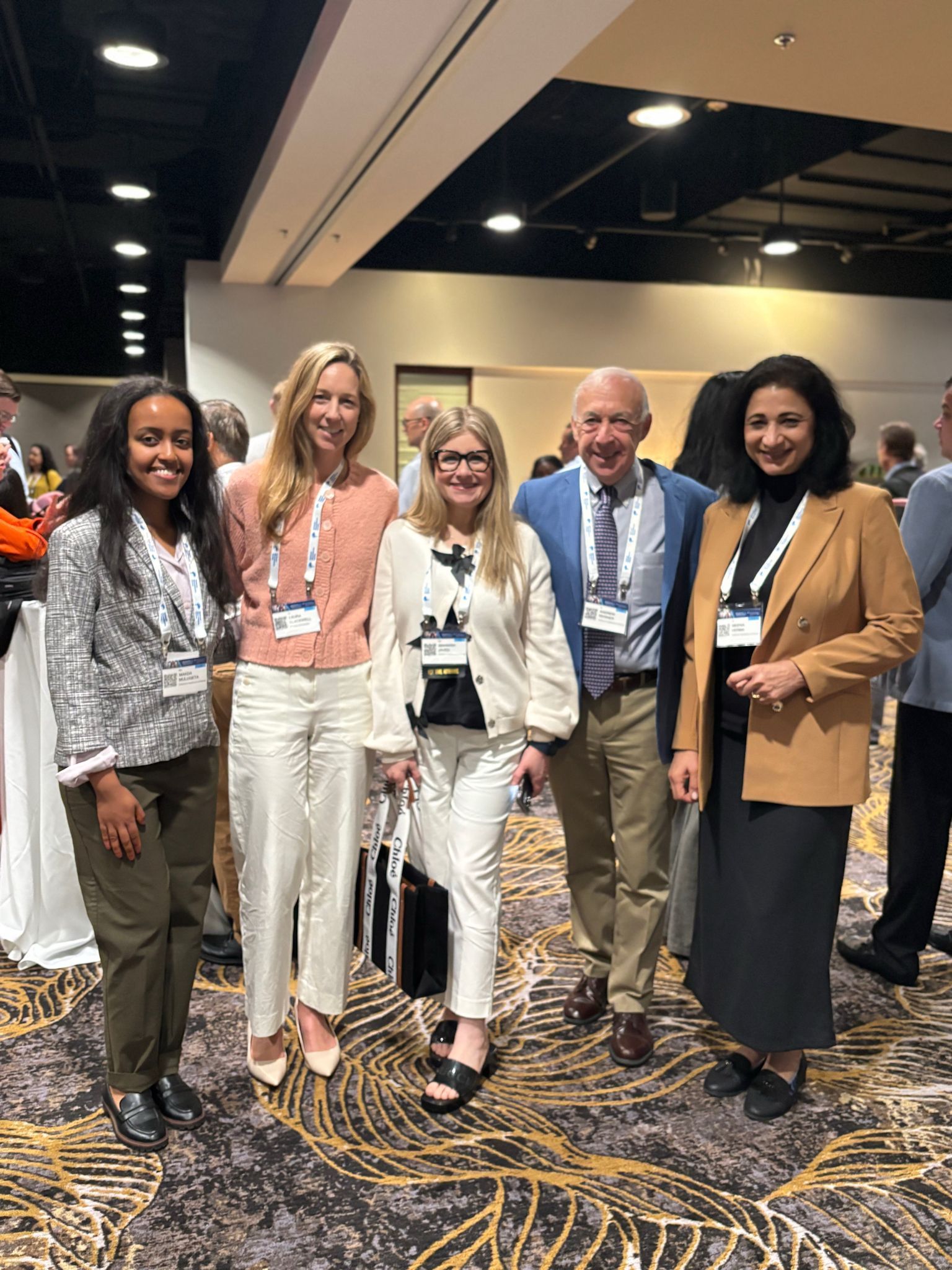 Five people posing indoors; smiling, wearing business attire, at a conference with patterned carpet.