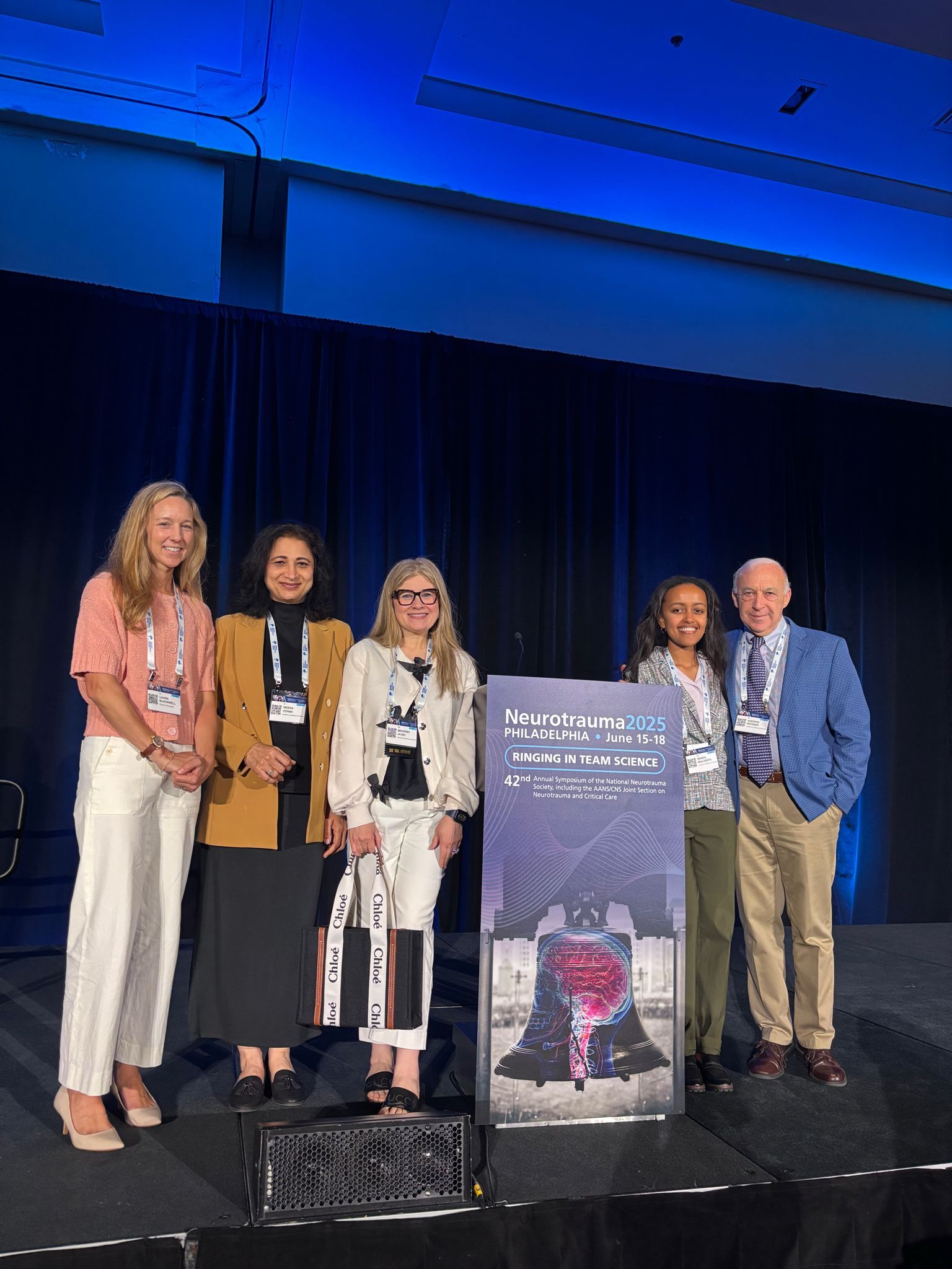 Six people standing onstage, near a sign, against a blue curtain.