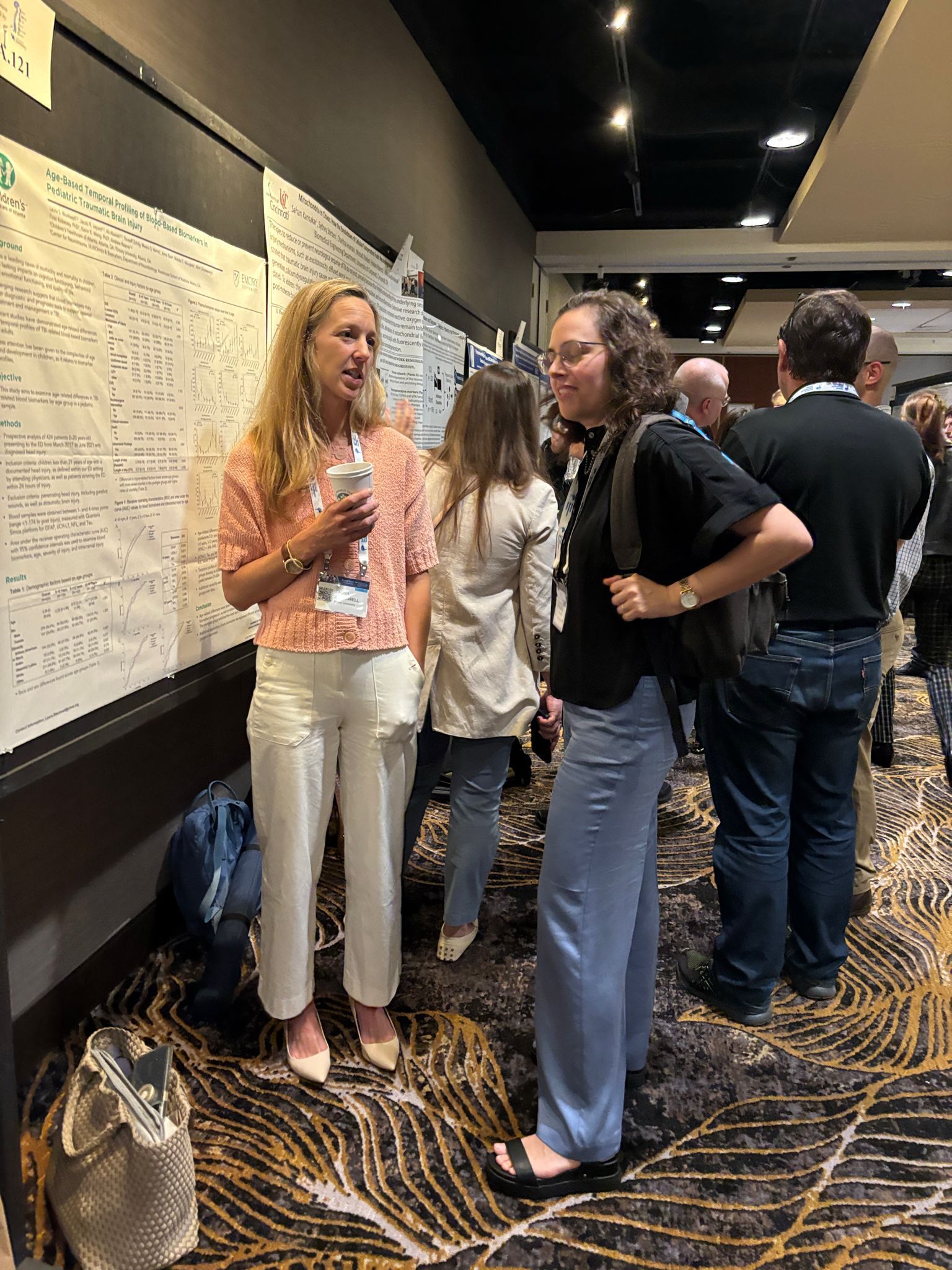 Two women converse near a poster board at a conference, surrounded by other attendees.