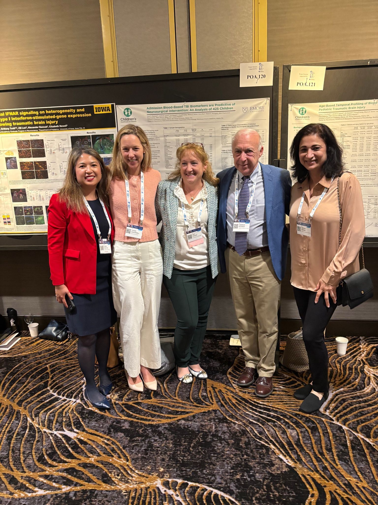 Five people pose in front of a conference poster. Three women and two men. Carpet is black with gold patterns.