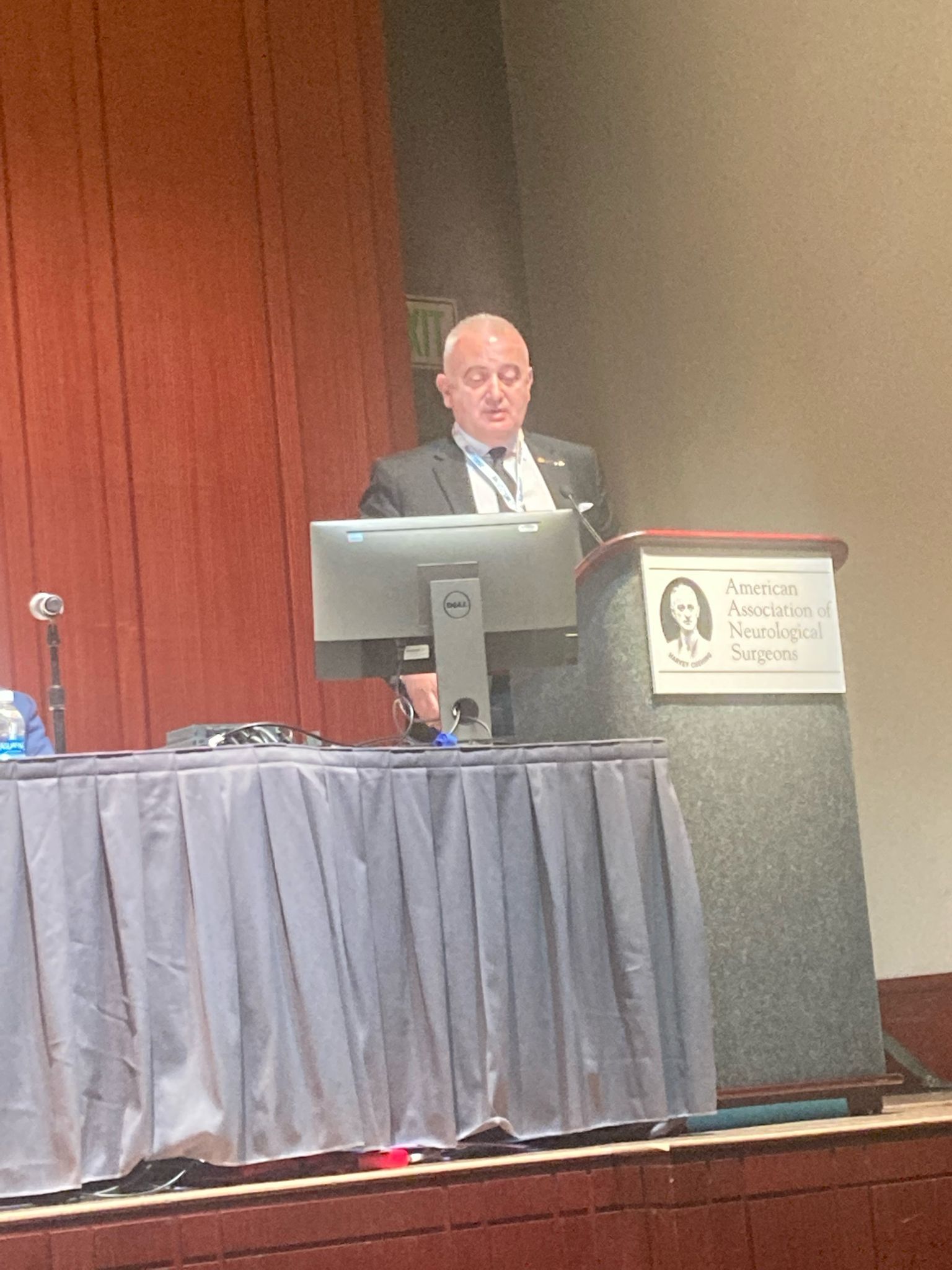 Man in suit speaking at a podium during a conference. Red backdrop, computer monitor, and microphone.