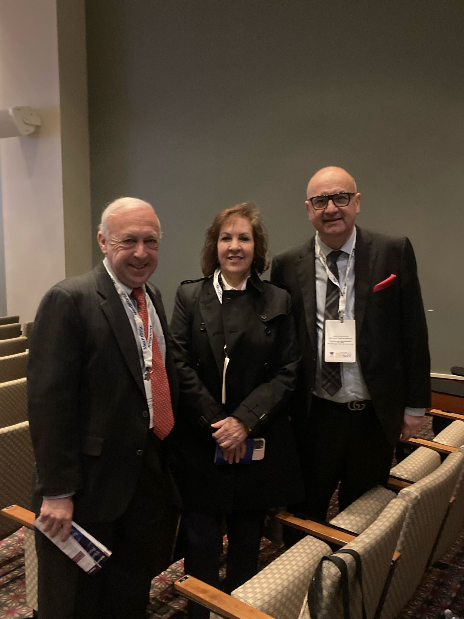 Three people standing near empty chairs in an auditorium. Woman in black jacket flanked by two men in suits.