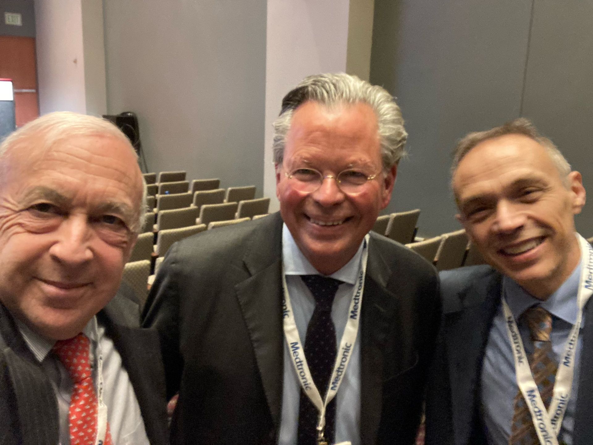Three men in suits smiling, indoors, in front of an auditorium.