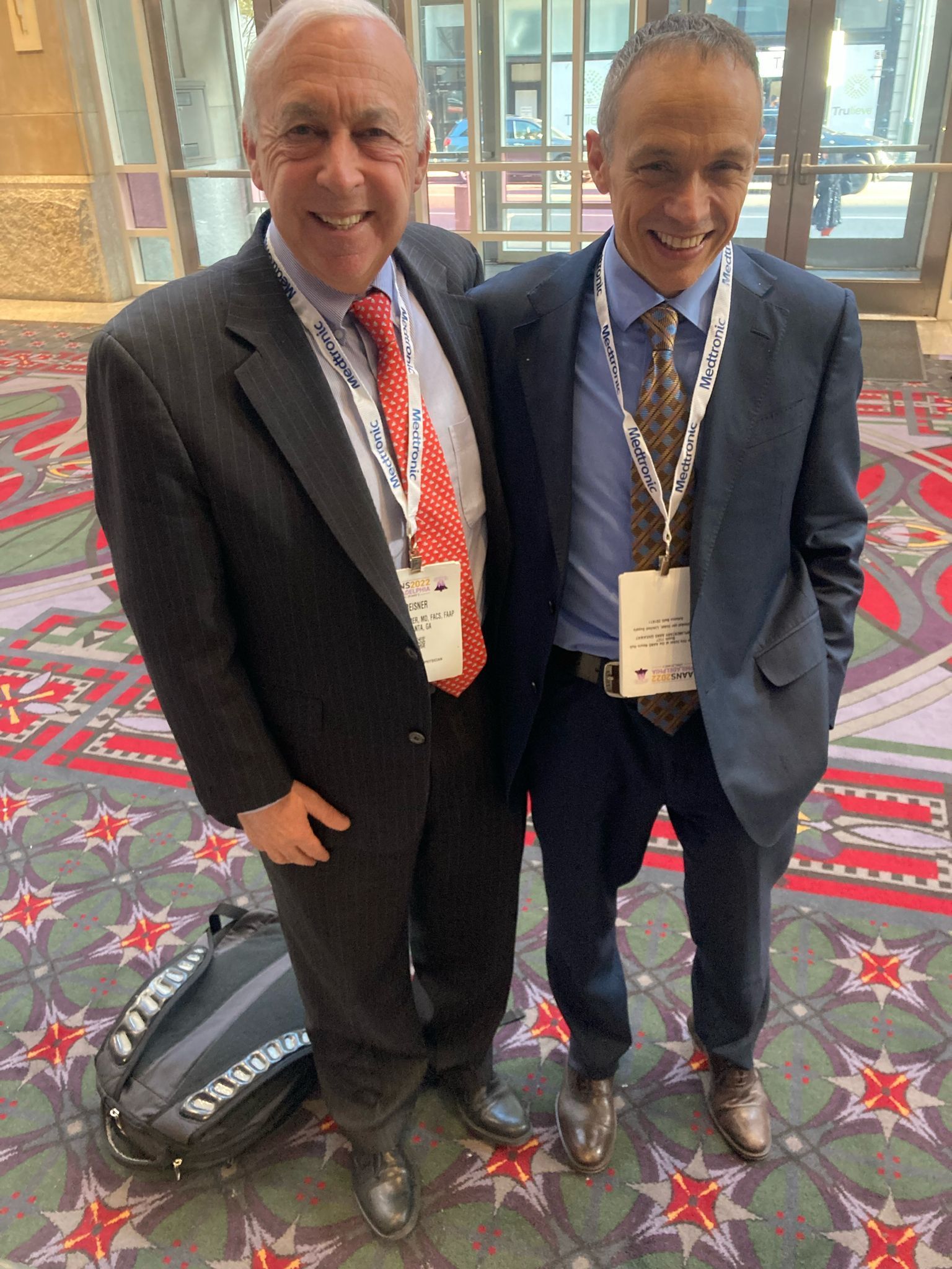 Two men in suits pose for a photo, smiling. One wears a red tie, the other a patterned tie. Inside a building.
