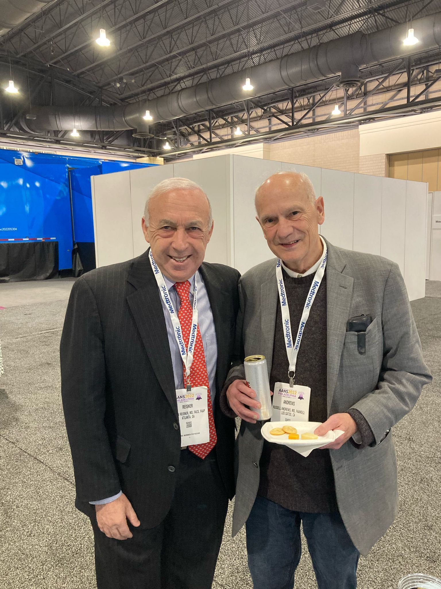 Two older men pose, one holds a snack, both wear lanyards, inside convention center with blue backdrop.