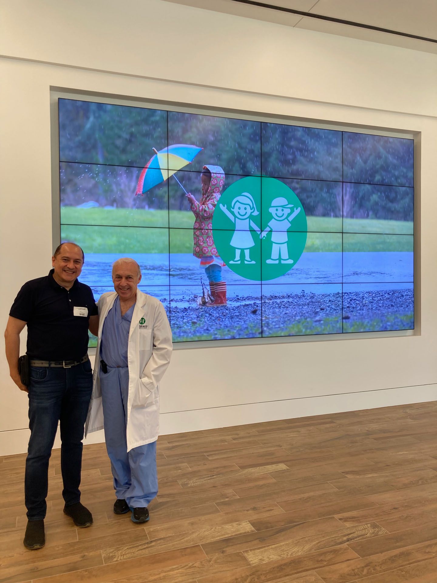 Two men pose in front of a large screen with a children's art display, inside a gallery.
