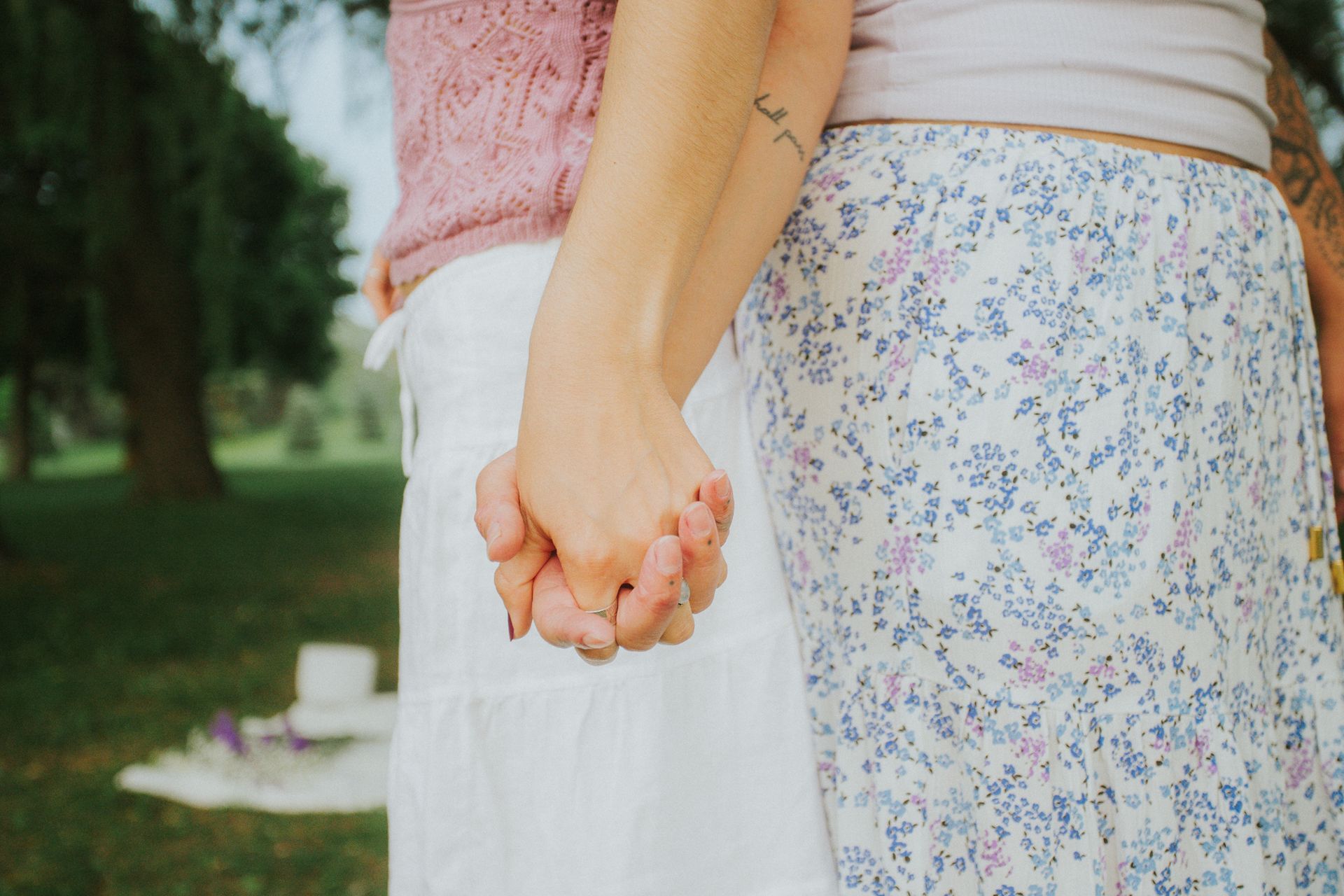 Two people holding hands in a park. One wears a pink sweater, the other a floral skirt.