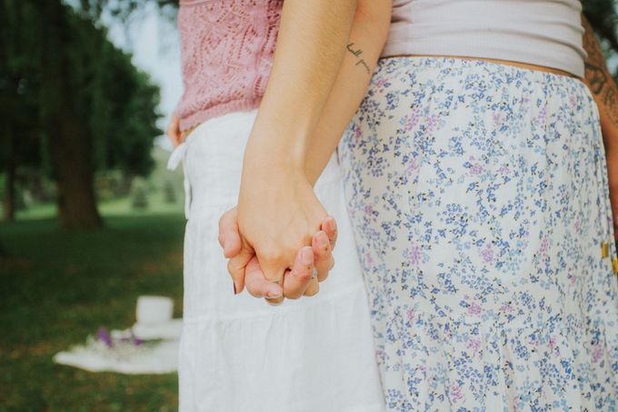 Two people holding hands in a park. One wears a pink sweater, the other a floral skirt.