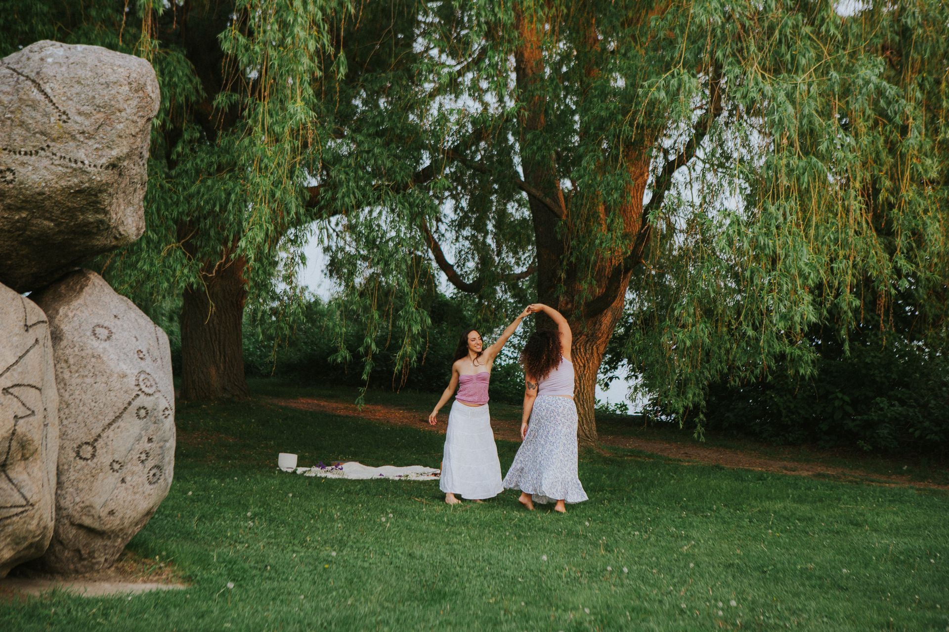 Two women dance under a willow tree, near large stones; a picnic blanket is on the grass.