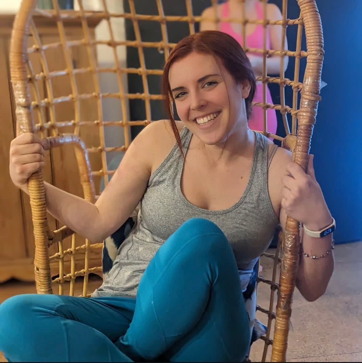 Woman with red hair smiles, sits in a rattan chair, wearing gray tank top and blue leggings.