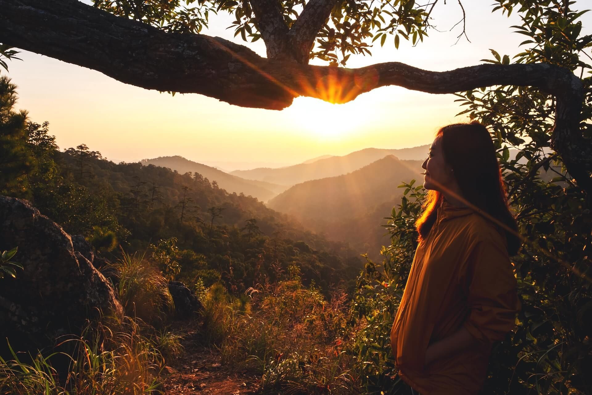 Woman in orange hoodie watches sunset over layered mountain range.