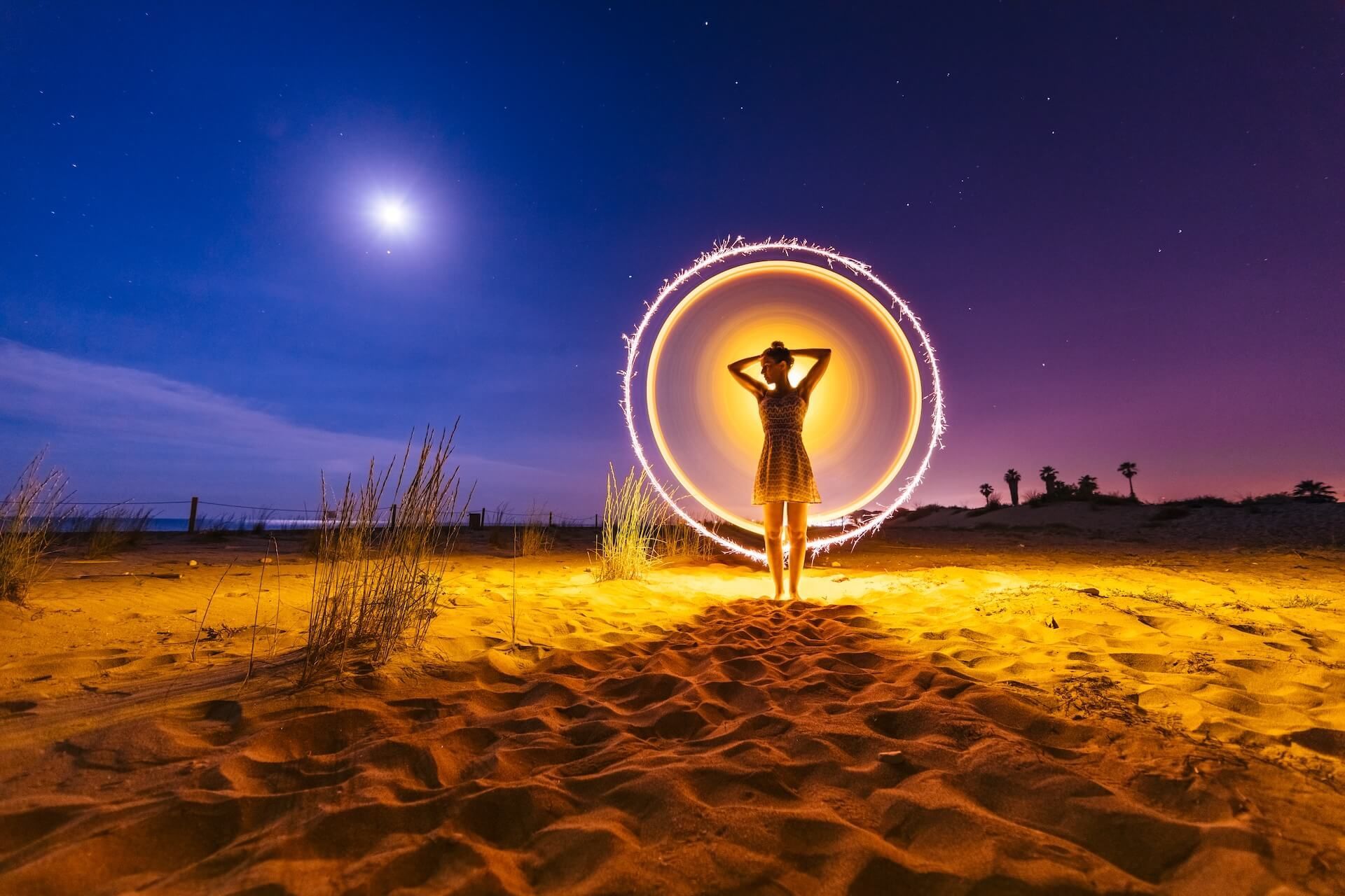 Woman silhouetted within a glowing circle of light, hands on head, desert setting, night sky with moon and stars.