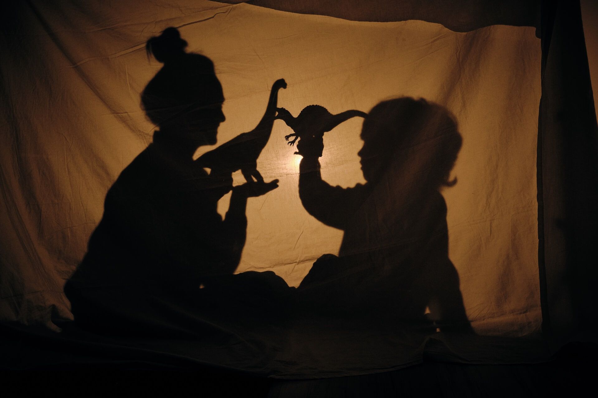 Silhouetted figures of a person and child playing with dinosaur toys, projected on a white sheet.