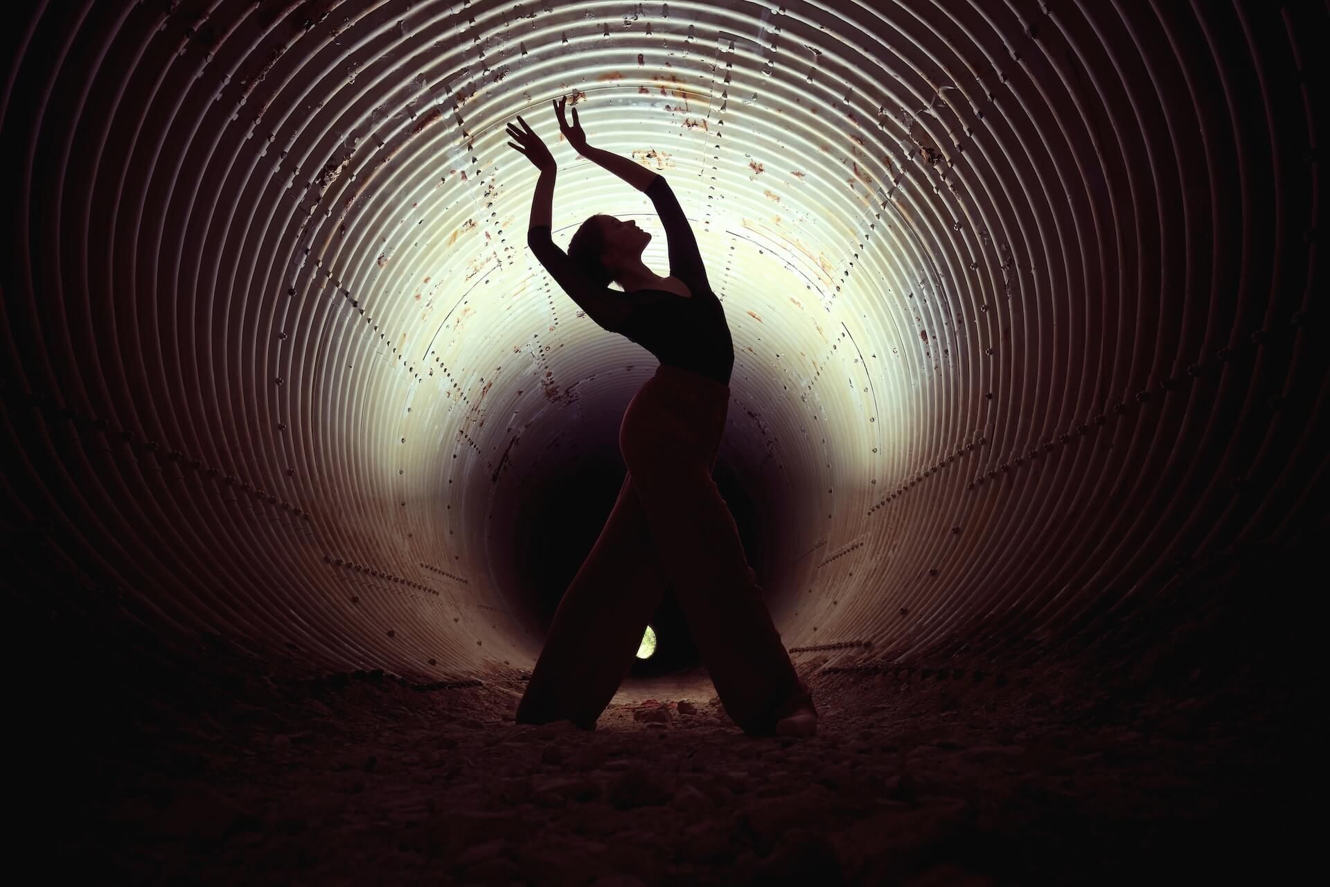 Dancer in silhouette arches back with arms raised inside a dark, corrugated tunnel lit from the end.