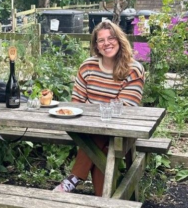 Woman in glasses sits at a picnic table in a garden, smiling. She wears a striped sweater and has a plate of food.