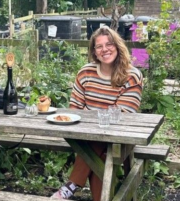 Woman with glasses smiles at a picnic table in a garden, with food and drinks.